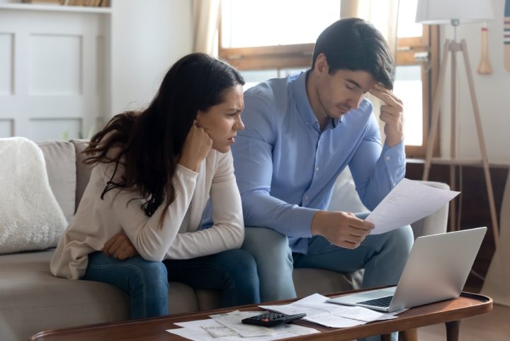 Falling behind with rent. Concerned worried young married couple sitting on sofa at home office studying paper letters from bank informing about debt bankruptcy. Financial loss unprofitable investment