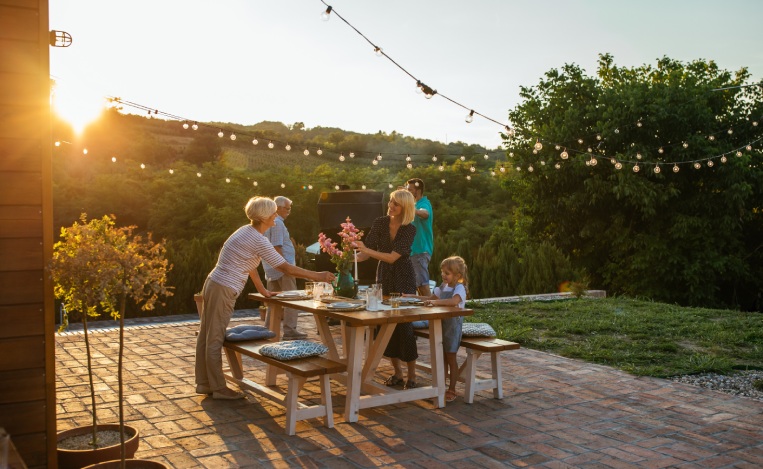 Whole family participates in the organization of dinner. Three female members setting dining table outdoors and male members barbecuing near them